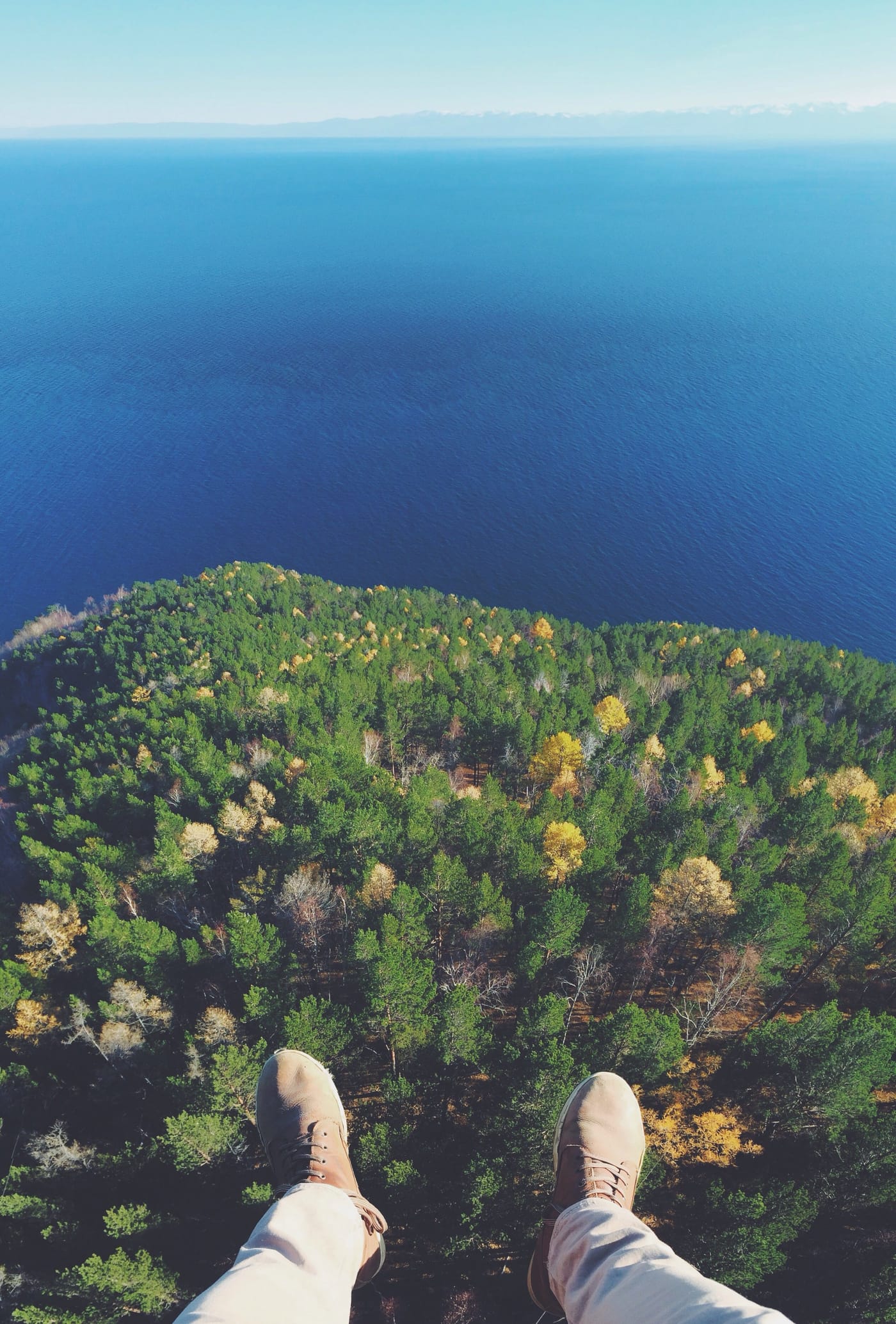 Looking down past a pair of boots at a forested headland and open blue water far below